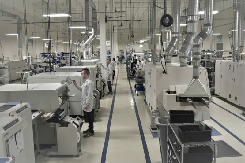 Technicians in white lab coats working amidst rows of large manufacturing equipment in a clean, modern electronic manufacturing facility.