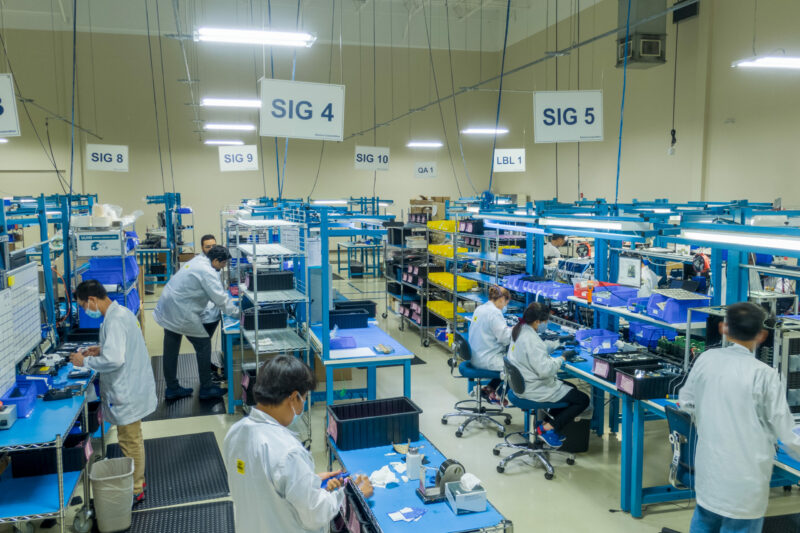 A busy electronic manufacturing services facility with workers in lab coats focused on assembling and testing electronic components at various workstations.
