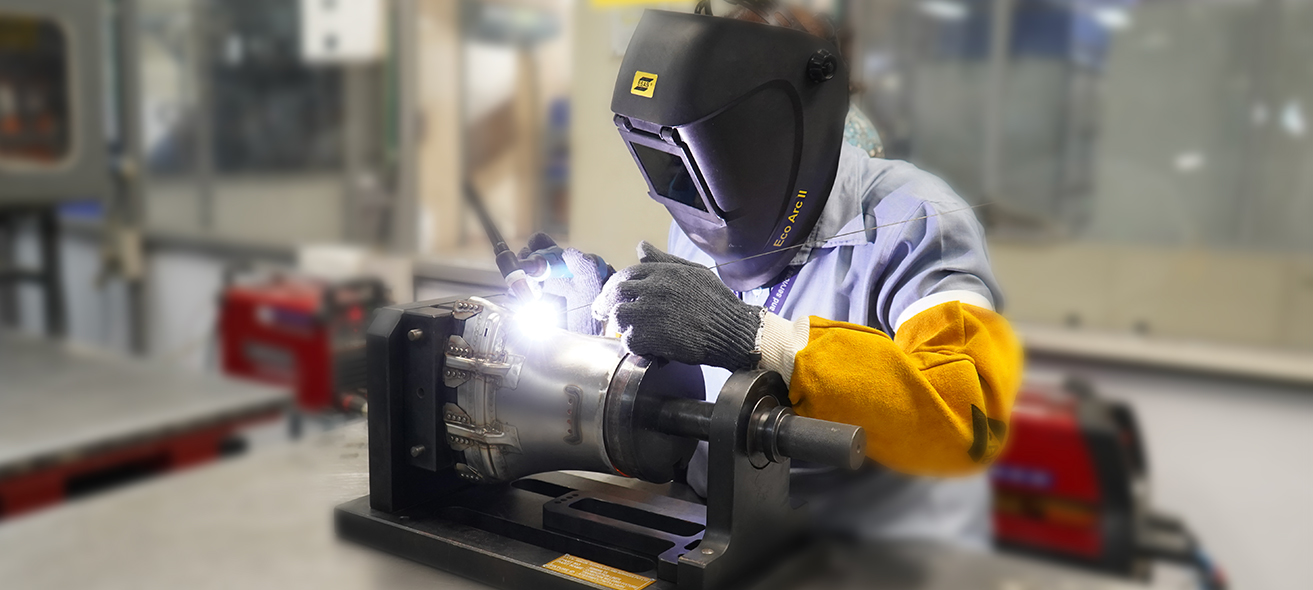 A welder in protective gear is performing precision welding on metal equipment in an industrial workshop, with bright welding light visible.