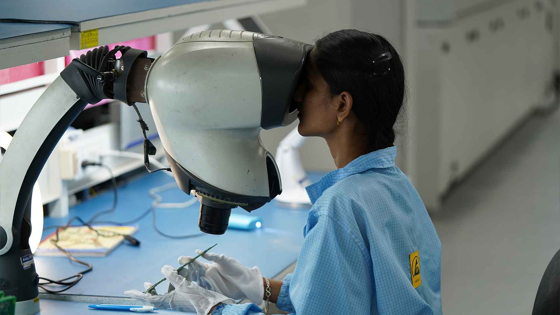 A technician in a light blue lab coat is closely examining a component through a large, professional microscope in an electronic manufacturing service lab.